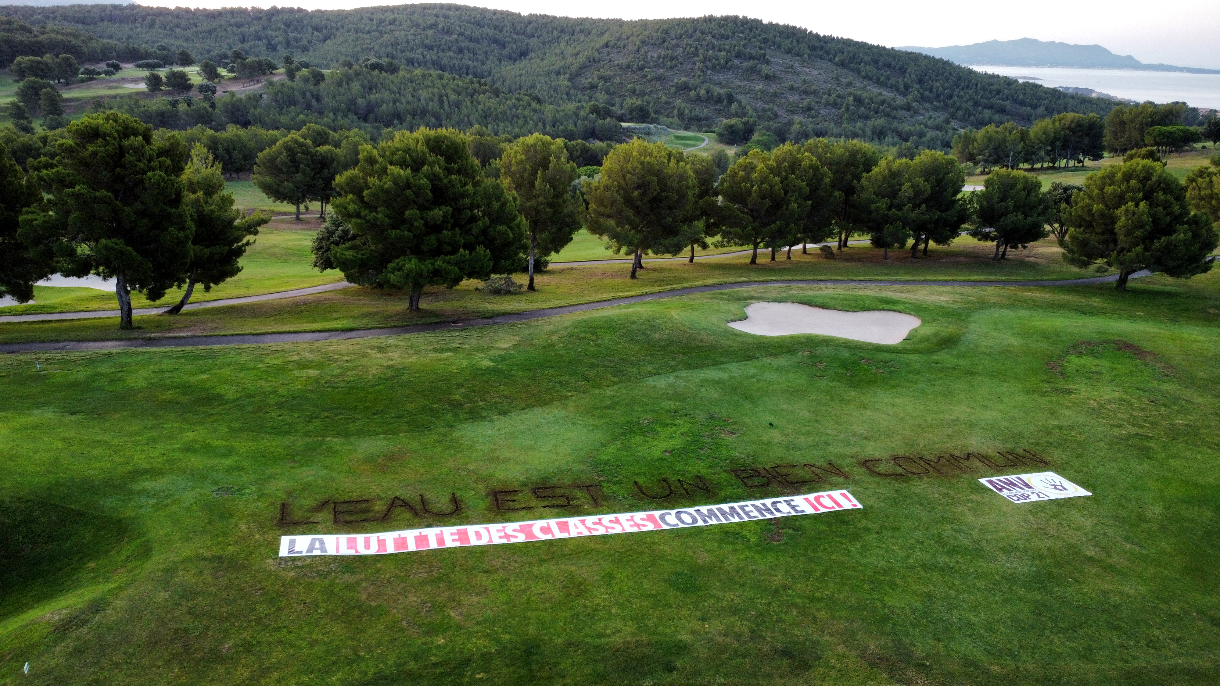 Golf vu de haut avec un message creusé dans la terre "L'eau est un bien commun" et une banderole "La lutte des classes commence ici"