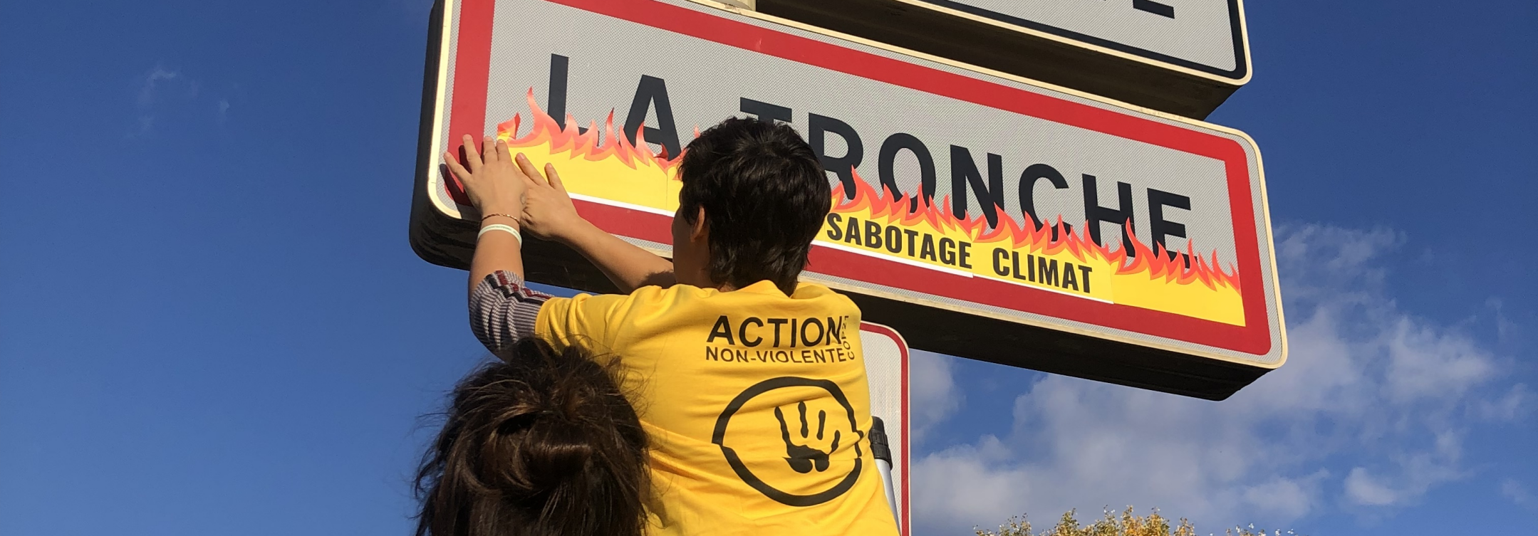 Une personne d'ANV-COP21 de dos colle des stickers flammes sur un panneau d'entrée de commune de 
